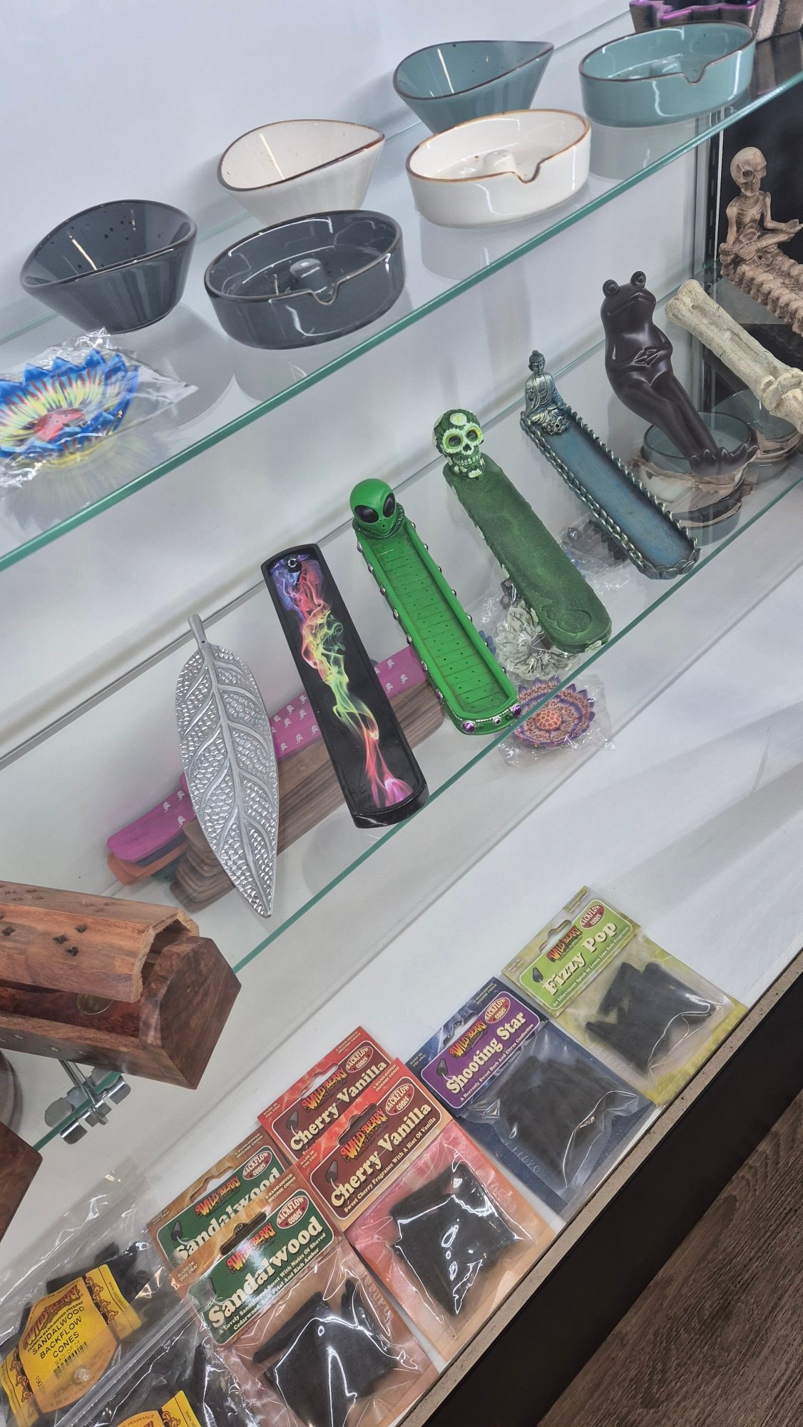 A glass shelf inside the shop: ceramic ashtrays on top, a row of incense holders below, an alien, a skull and a silver leaf burner, with packets of backflow incense cones along the counter.